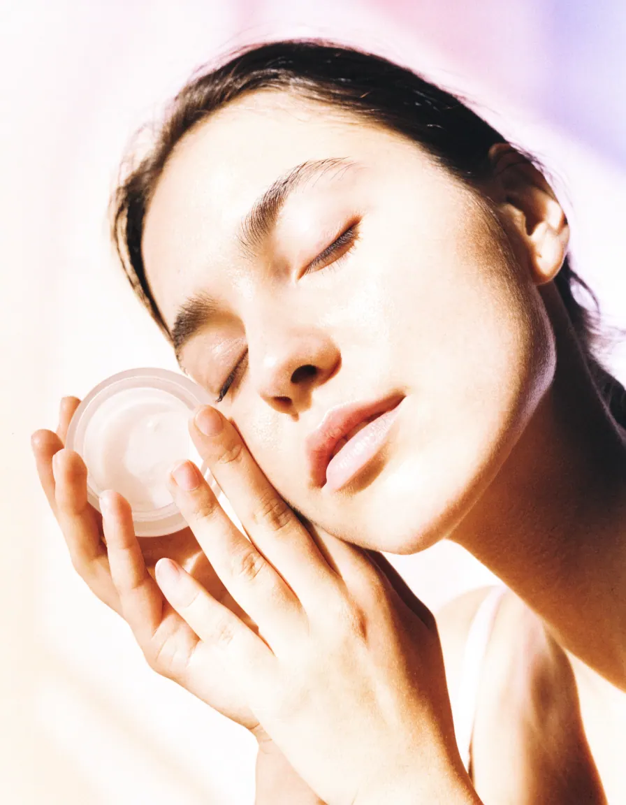 A woman with her eyes closed holding a small frosted cream jar beside her cheek in soft pink light