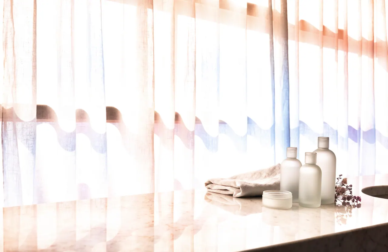 A bathroom vanity with frosted skincare bottles, a linen towel and dried flowers in morning light